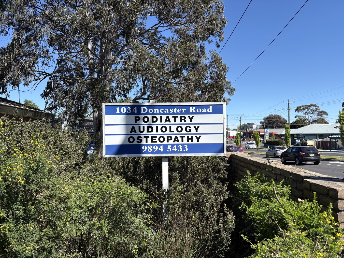 The blue and white sign at the street front of 1034 Doncaster Road, listing podiatry, audiology and osteopathy, with the number 9894 5433